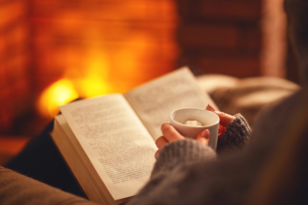 A reader curled up by a glowing fireplace, holding a cup of hot cocoa while reading a romance novel—capturing the warmth and comfort of cozy season romance.