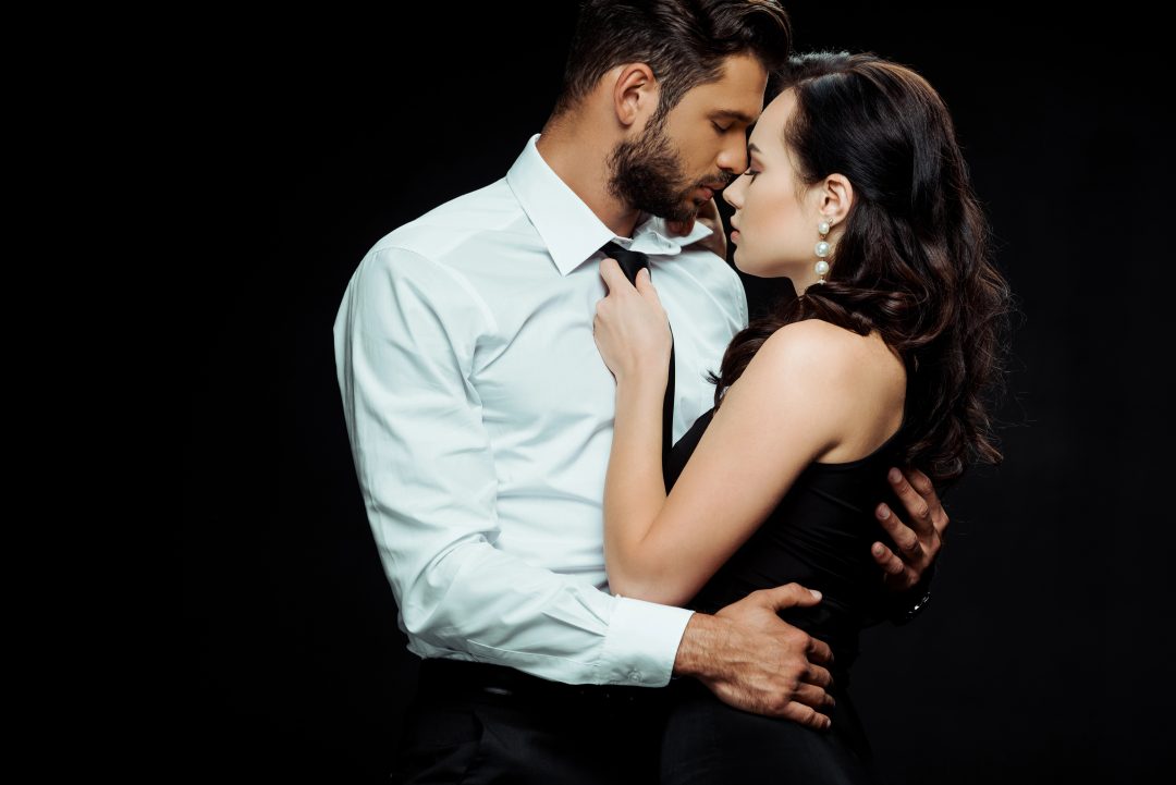 A man in a white dress shirt holds a woman close as they lean in for a slow, intimate kiss, capturing the sensual tension of a slow burn romance.